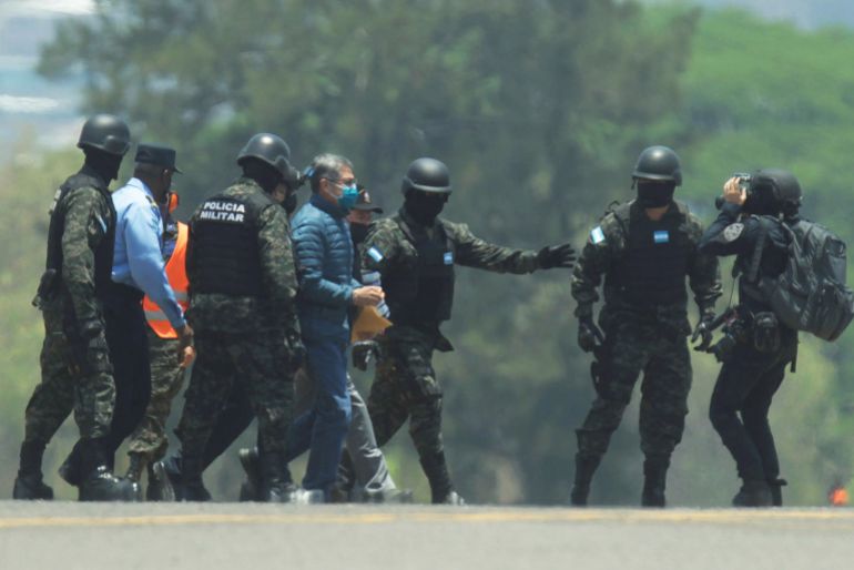 Национальная полиция Гондураса в касках и бронежилетах сопровождает бывшего президента Хуана Орландо Эрнандеса через дорогу.